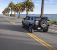 2024 GMC HUMMER EV SUV drives down a coastal road with palm trees.