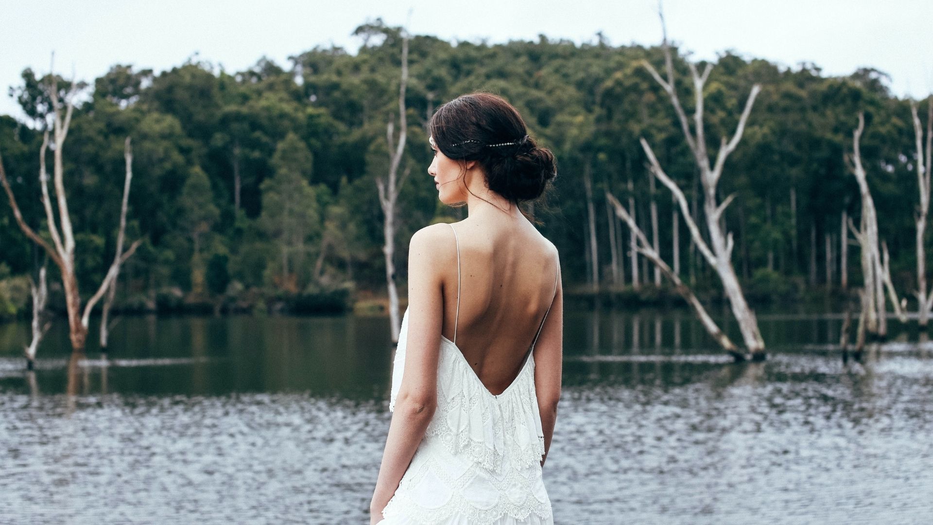 Une femme en robe de mariée dos-nu se tient devant une étendue d'eau