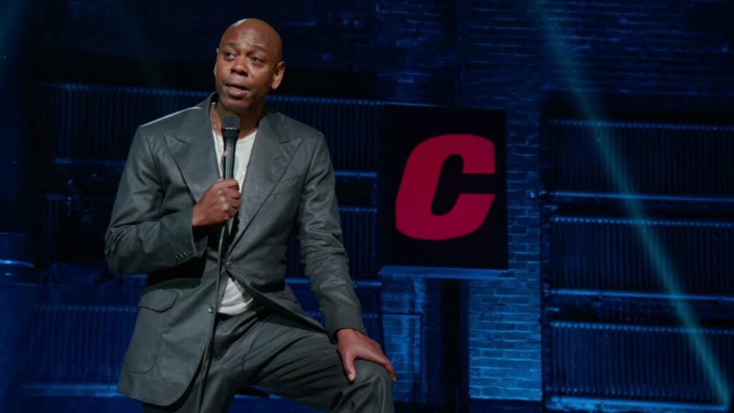 Le comédien de stand-up Dave Chappelle est debout sur scène, micro à la main. Le comédien de stand-up Dave Chappelle est debout sur scène, micro à la main.