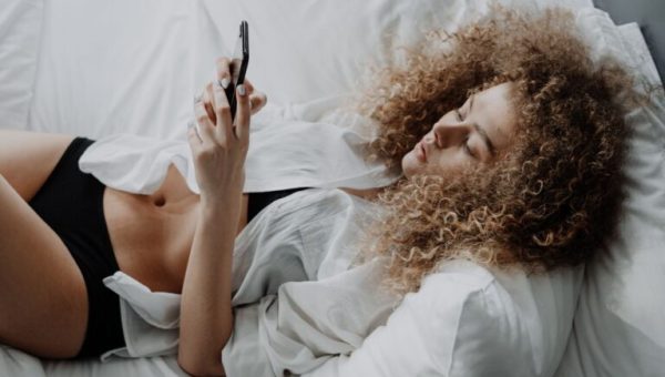 Une femme dénudée allongée dans son lit, en train de consulter son téléphone portable