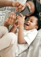 Une mère et son enfant, allongés dans un lit, en train de regarder un écran de téléphone portable et de rire // Source : Getty Images Signature