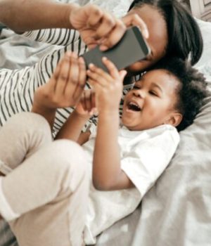 Une mère et son enfant, allongés dans un lit, en train de regarder un écran de téléphone portable et de rire // Source : Getty Images Signature