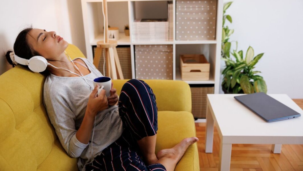 Une femme assise dans son salon en train de boire du café et d'écouter de la musique ou un podcast avec un casque audio, les yeux fermés, l'air détendu // Source : bojanstory de Getty Images Signature