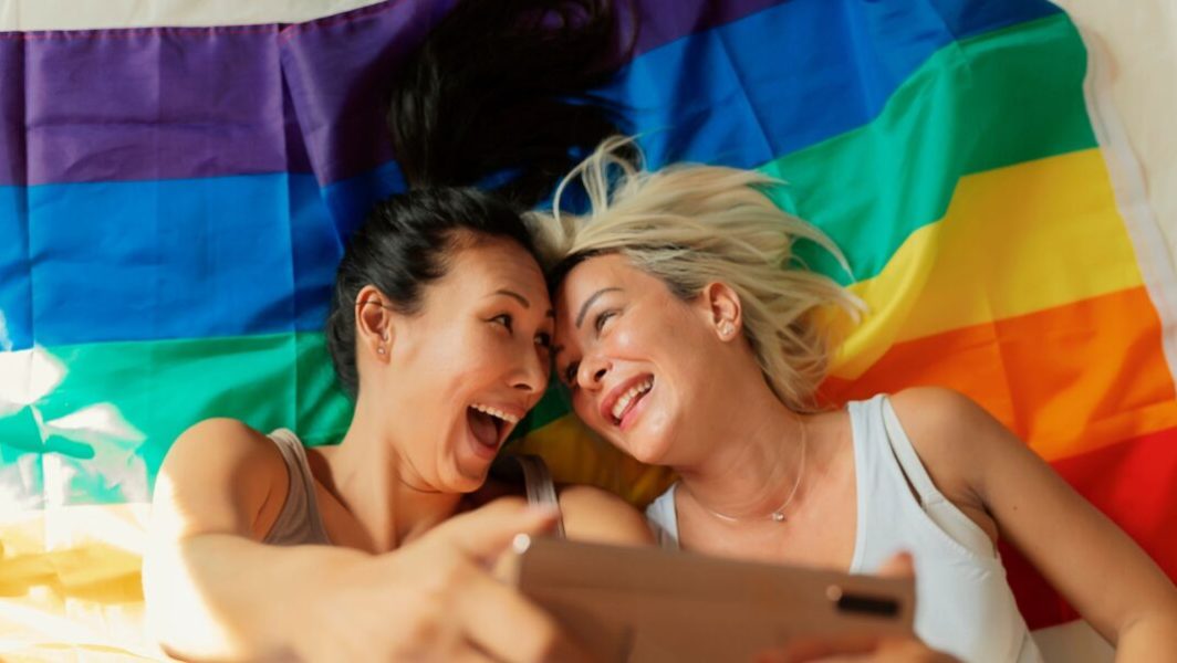 Deux femmes lesbiennes en train de faire un selfie devant un drapeau LGBT+ // Source : PixelsEffect de Getty Images Signature