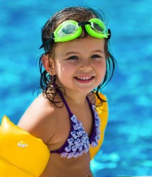 Une petite fille avec des lunettes de piscine fluo, un maillot de bain bleu et des brassards flotteurs jaunes devant une étendue d'eau // Source : Getty Images Signature