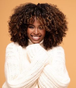 Une femme aux cheveux frisés qui sourit en regardant l'objectif, habillée d'un gros pull douillé col roulé blanc crème sur un fond beige.jpg // Source : NeonShot de Getty Images