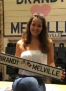 Une femme pose avec un panneau au logo de la marque Brandy Melville, au sein d'une boutique // Source : Capture d'écran YouTube de la bande-annonce du documentaire "Brandy Hellville & The Cult of Fast Fashion"