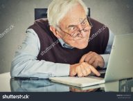 stock-photo-elderly-old-man-using-computer-sitting-at-table-isolated-on-grey-wall-background-senior-people-and-252858463