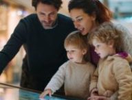 Un couple et ses enfants regardent une borne intéractive dans un centre commercial pour se repérer // Source : Midjourney - image générée par IA
